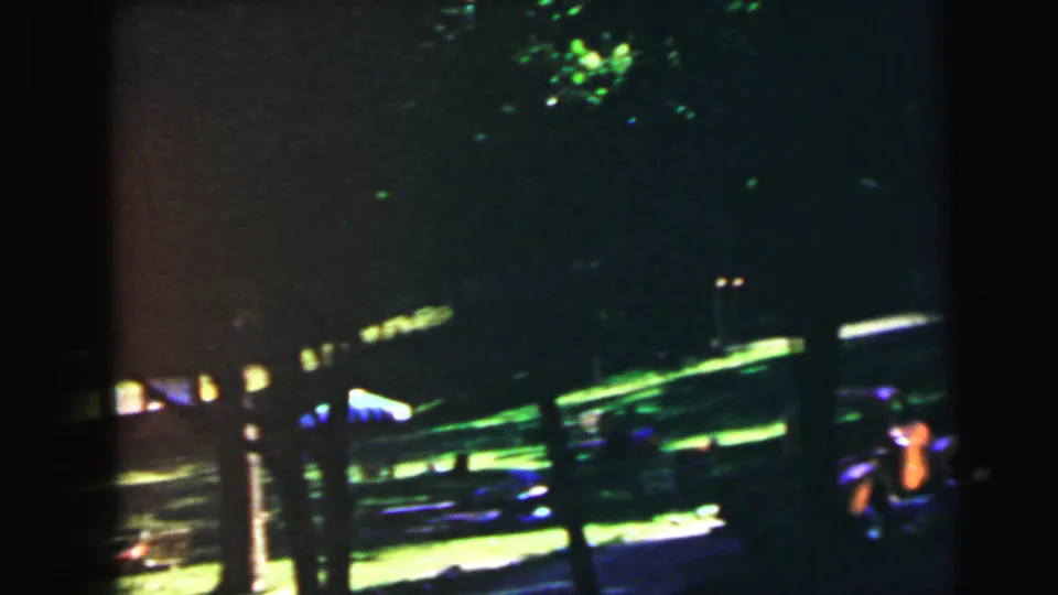 8mm home movie frame of a summer yard shaded by mature trees, with sunlight breaking through onto green grass and a small colourful object at the far right edge; suburban New Jersey, circa 1964.