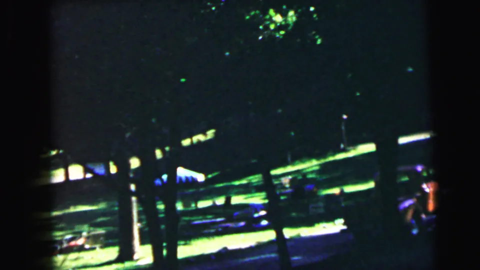 Heavily underexposed 8mm frame of a shaded suburban yard; tree trunks and faint lawn visible beneath dense canopy with minimal light penetration; suburban New Jersey, circa 1964.