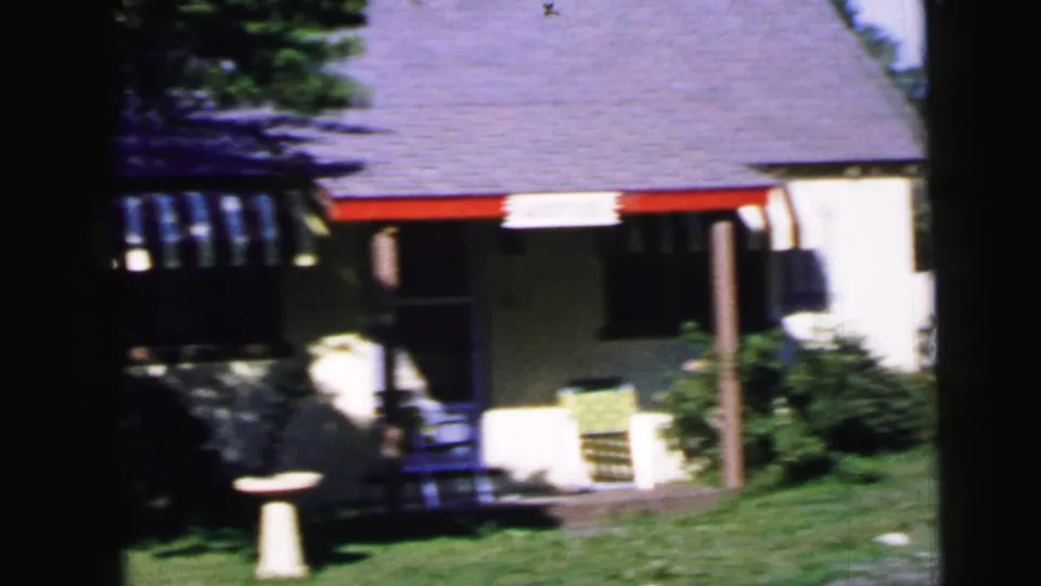 Clear 8mm frame of a mid-century suburban house exterior with gray shingle roof, white siding, blue-and-white striped porch awning, outdoor seating, and a birdbath in the foreground; consistent with Camden, New Jersey, 1964.