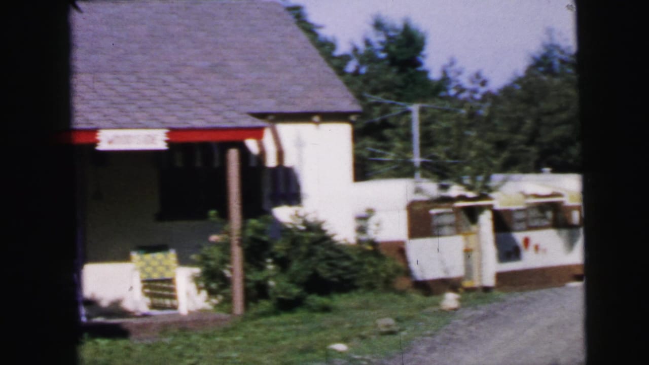 8mm home movie frame circa 1964 showing a modest suburban home exterior with red porch trim, a white-and-brown travel trailer, lawn chair, and summer trees, described as Camden, New Jersey.