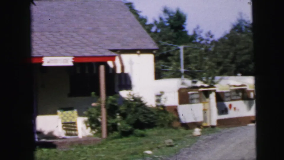 8mm home movie frame circa 1964 showing a modest suburban home exterior with red porch trim, a white-and-brown travel trailer, lawn chair, and summer trees, described as Camden, New Jersey.