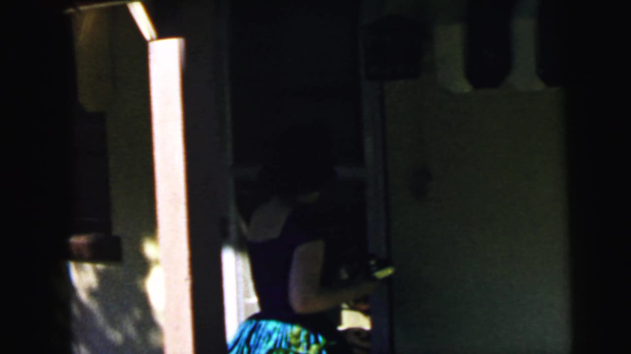 8mm home movie frame circa 1964 showing a woman in a blue-green patterned dress entering a darkened interior doorway, backlit by bright sunlight.