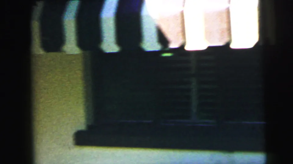 Dark underexposed 8mm transitional frame circa 1964, possibly showing a porch or covered entry structure with vertical posts as the camera moves from exterior to interior.