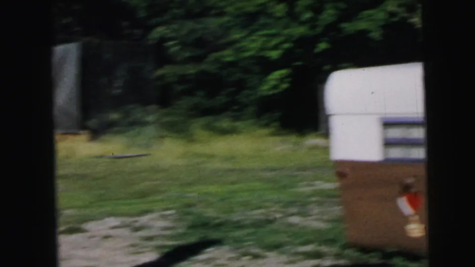 8mm home movie frame circa 1964 showing a grassy suburban yard with dense summer trees and a white-and-brown travel trailer partially visible at frame right, with a red or orange object near its base.