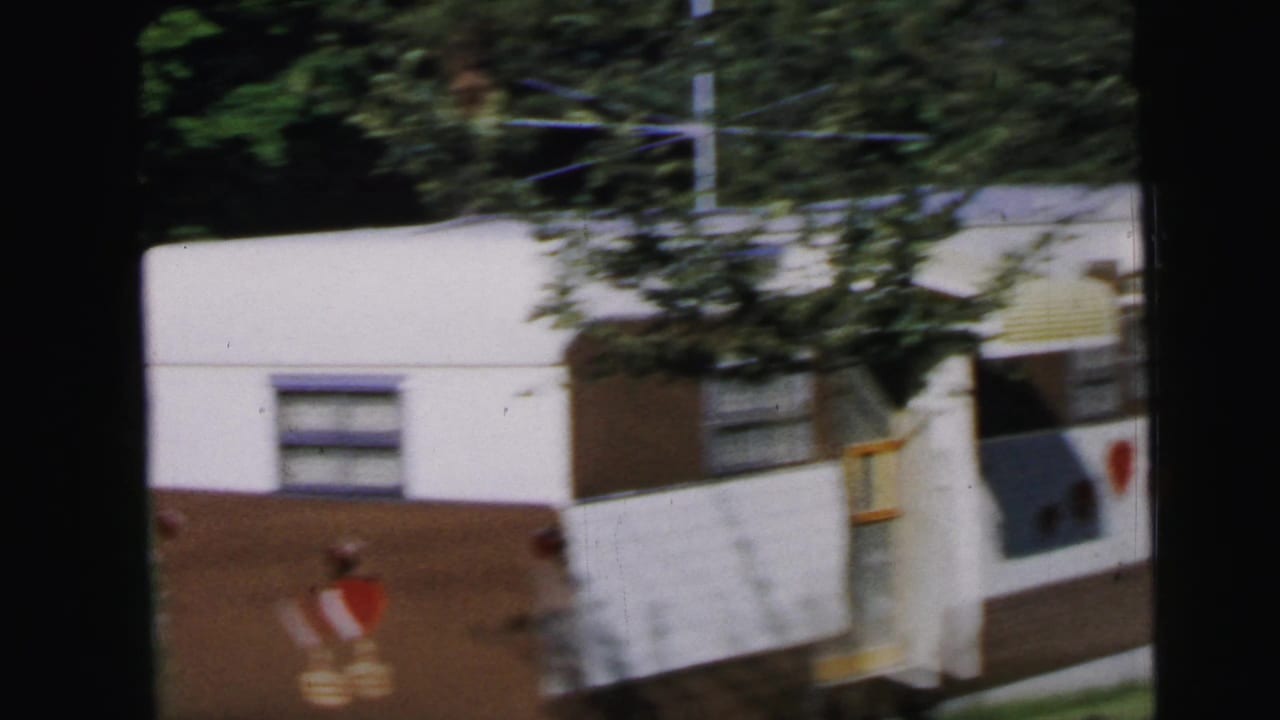 Close shot of a white-and-brown mid-1960s travel trailer with yellow-striped awning, surrounded by summer-green trees, from an 8mm home movie circa 1964 in Camden, New Jersey.