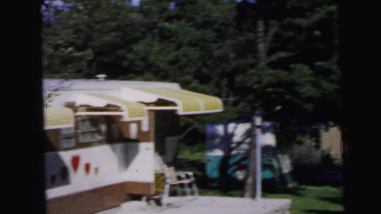 White and brown travel trailer with yellow-striped awning, folding chair alongside, at a sunny wooded campsite.