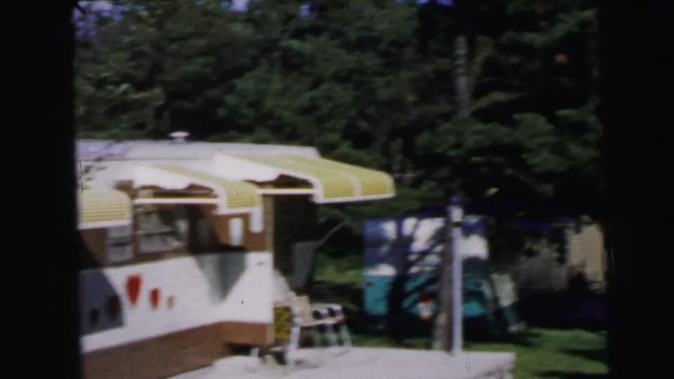 Mid-1960s travel trailer with yellow-striped awning and folding chair outside, in a wooded New Jersey campground, from 8mm home movie footage circa 1964.