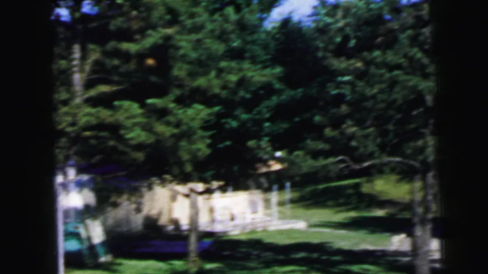 Wide view of a wooded summer campground with a camper visible through the trees, from an 8mm home movie shot in New Jersey circa 1964.