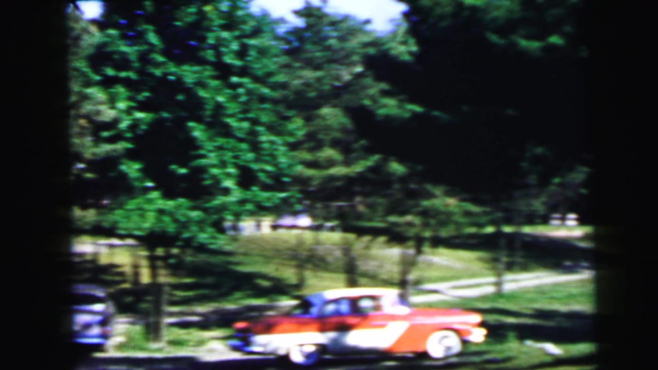 Red and white two-tone classic American car parked in a sunlit wooded campground; a second darker vehicle is partially visible at left.