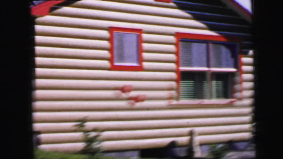 Camper trailer or cabin exterior with log-cabin-style tan siding and red window trim, a mid-century recreational vehicle style consistent with 1960s campsite use.