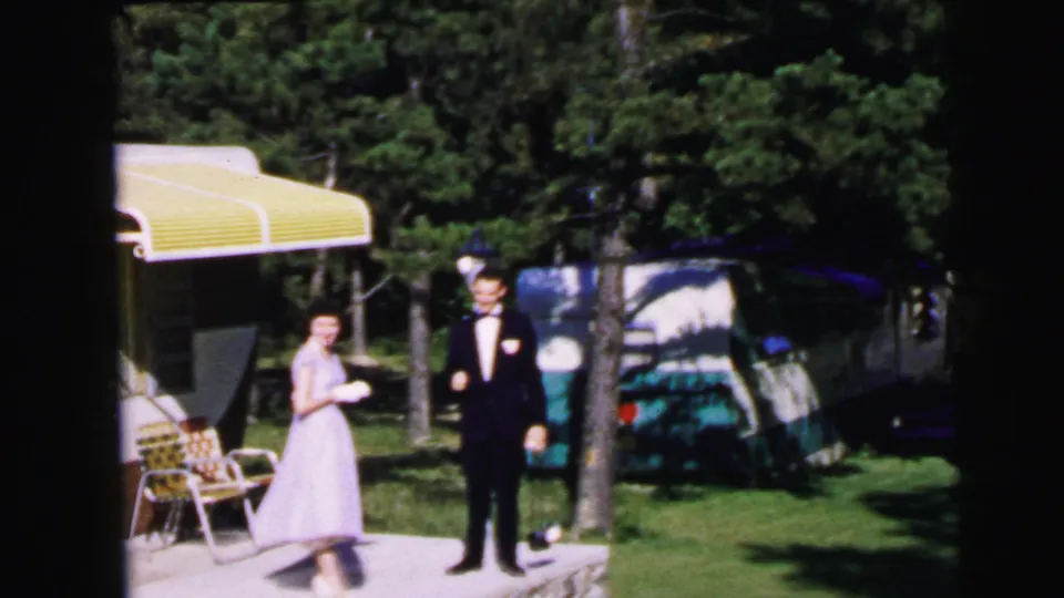 Wedding couple in pale gown and dark tuxedo at a wooded campsite, circa 1964, consistent with Camden NJ footage; yellow awning, folding chairs, and teal car behind them.