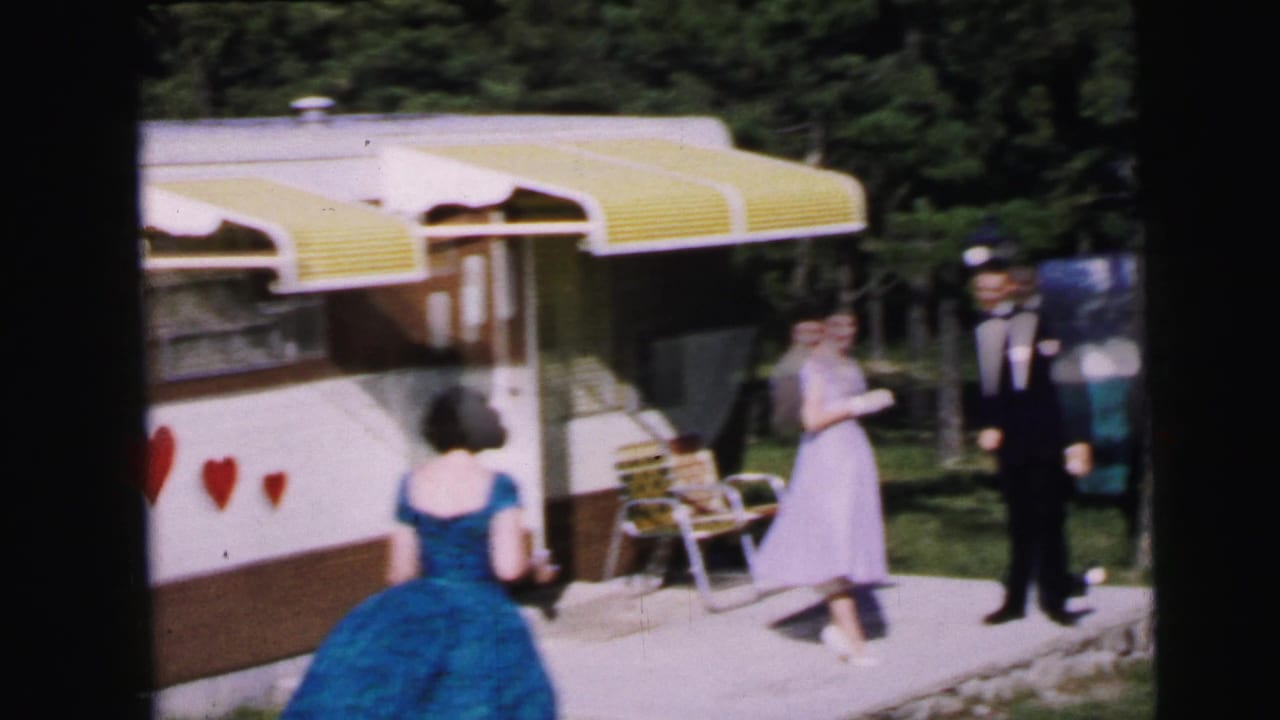 1964 home movie frame: two women in formal dresses and a man in a tuxedo gathered near a travel trailer with yellow awnings and red heart decorations, Camden, NJ.