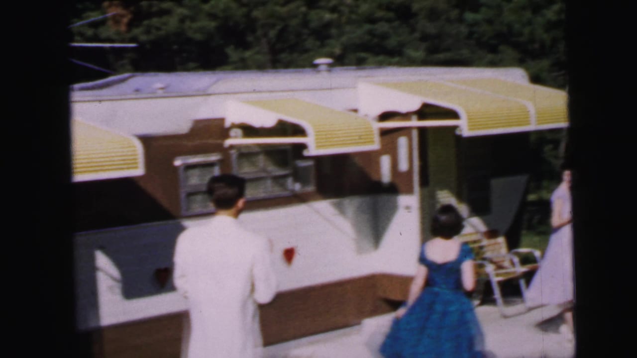 1964 home movie: two figures from behind — man in white jacket, woman in blue dress — before a travel trailer with yellow awnings and a red heart painted on its side, Camden, NJ.
