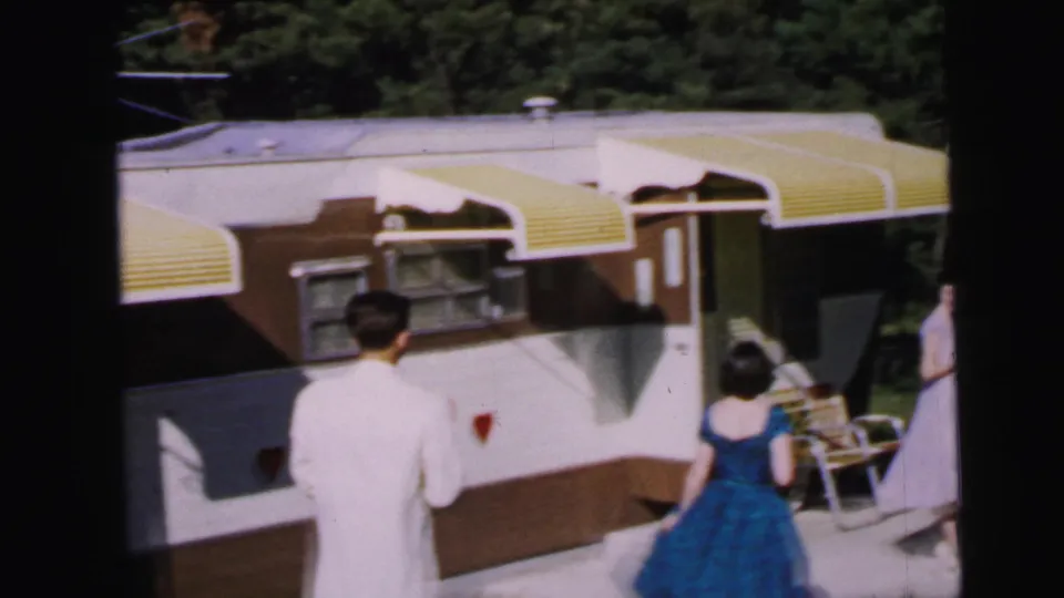 1964 home movie: two figures from behind — man in white jacket, woman in blue dress — before a travel trailer with yellow awnings and a red heart painted on its side, Camden, NJ.