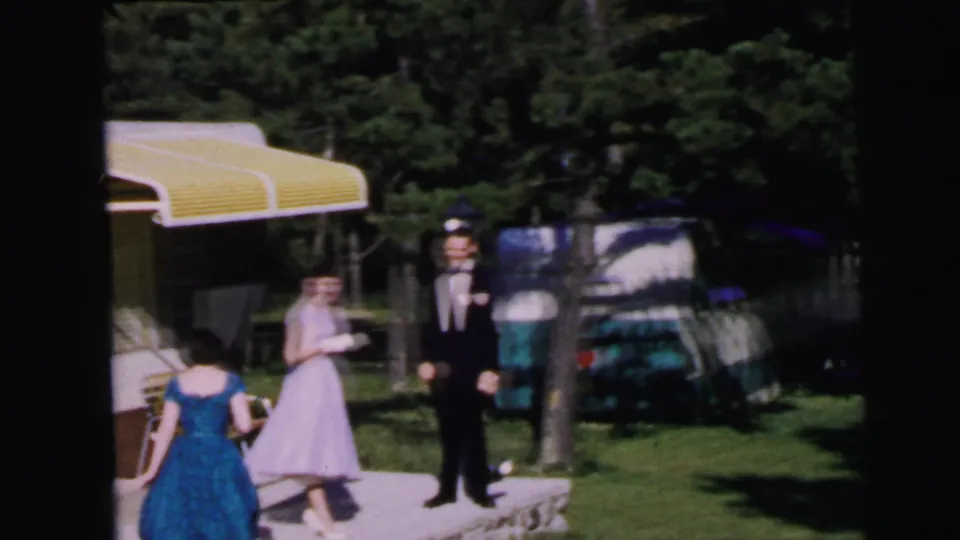 1964 home movie: three formally dressed figures — blue full-skirted dress, lavender gown, dark tuxedo — gathered beside a yellow-awninged travel trailer in a wooded campground setting, Camden, NJ.