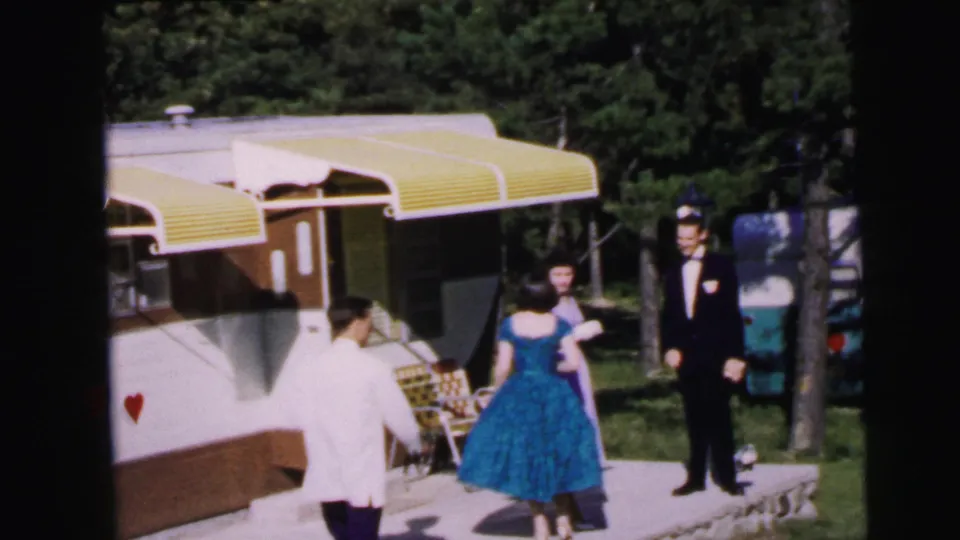 1964 home movie: man in white jacket, woman in blue full-skirted dress, and man in dark tuxedo gathered by a travel trailer with yellow awnings and a red heart motif on its side, Camden, NJ.