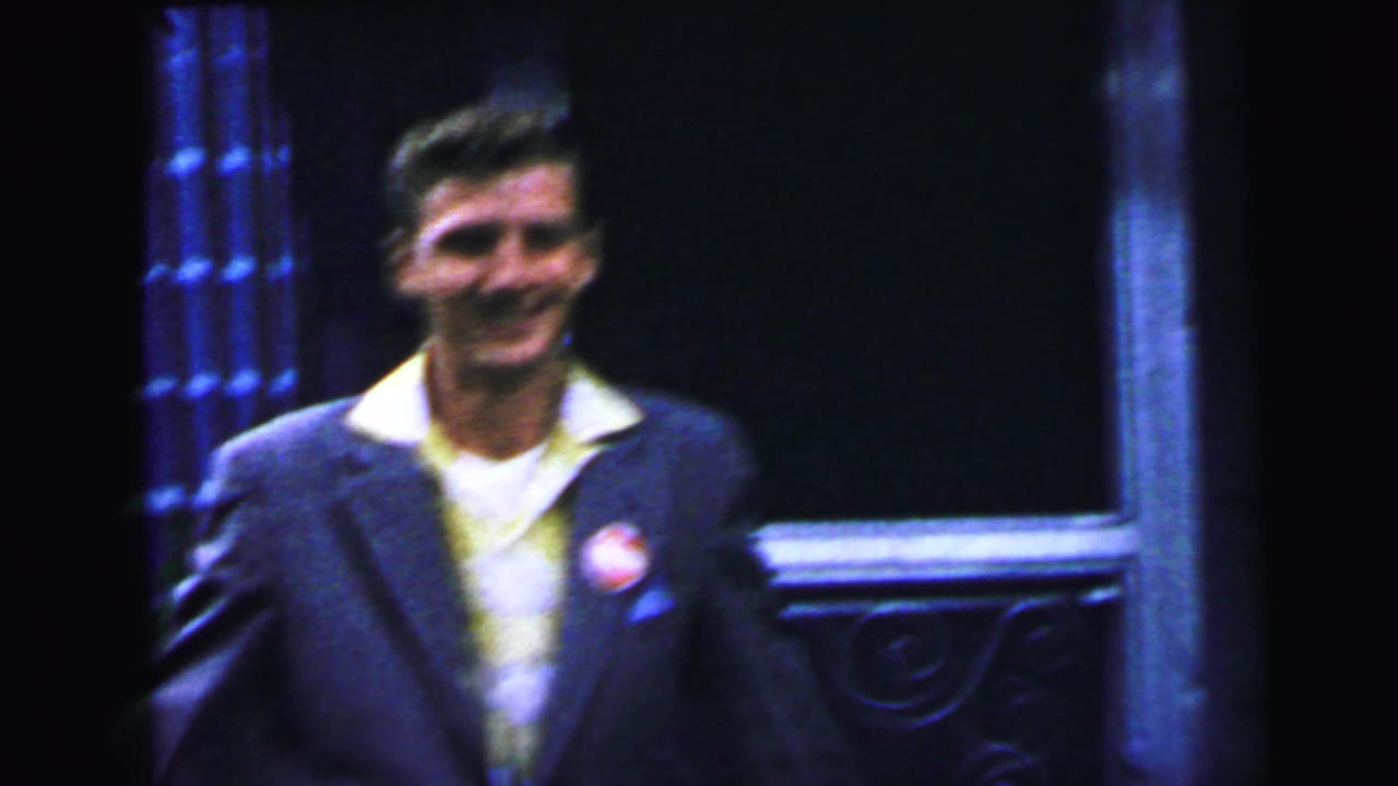 Young man in early-1960s narrow-lapel suit and yellow shirt smiling at camera, round red-and-white lapel button on left breast, standing before an ornate cast-iron doorway surround, Camden NJ area, 1964.