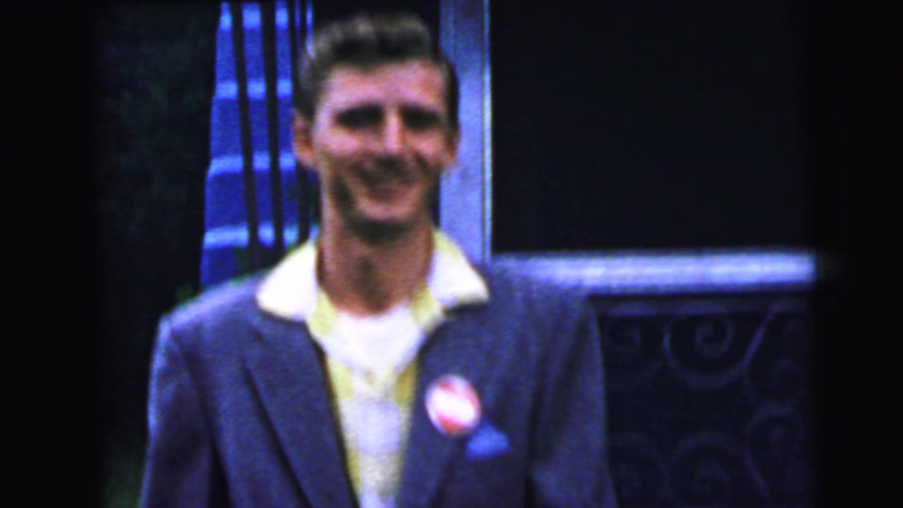 Young man in dark suit and yellow shirt smiling broadly, round lapel button visible, decorative ironwork surround to right.