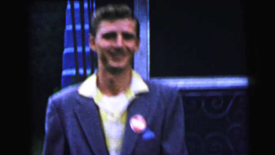 Young man in 1960s suit and open-collar yellow shirt smiling broadly at camera, round lapel button on left breast, ornate cast-iron door surround visible to right, consistent with Camden NJ, 1964.