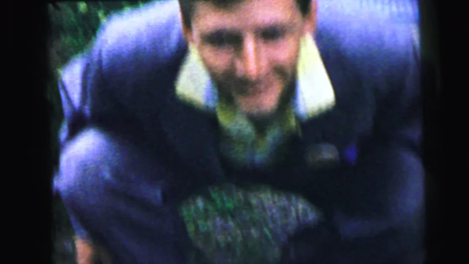 Young man in dark suit jacket leaning toward the camera with a broad grin, yellow shirt visible, green foliage in background — an informal home-movie gesture, possibly Camden NJ, 1964.