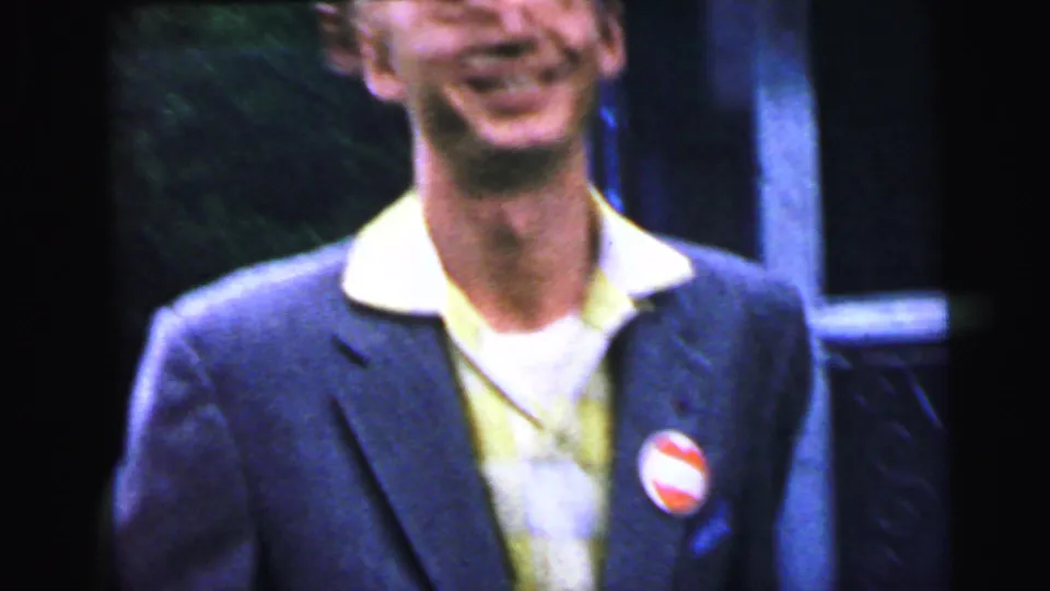 Young man in 1960s suit and yellow open-collar shirt laughing toward camera, round red-and-white lapel button clearly visible on left breast, ornate cast-iron surround to right, consistent with Camden NJ, 1964.