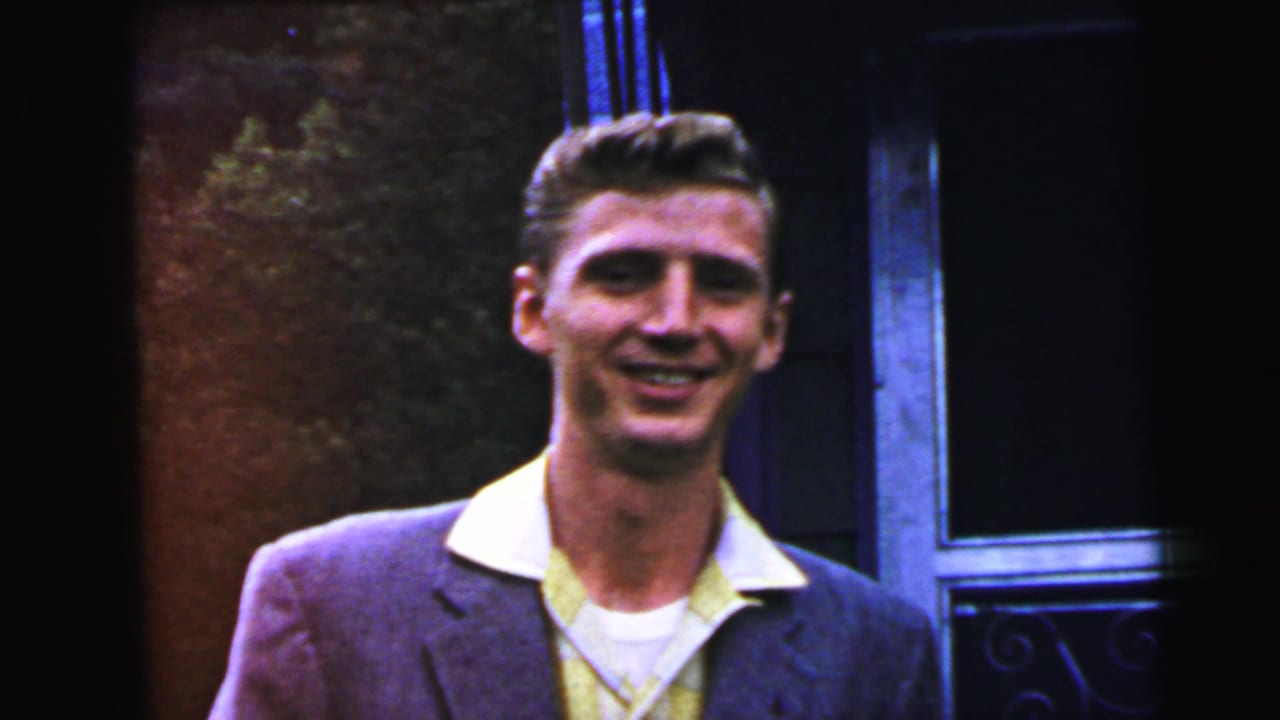 Young adult man in early-1960s narrow-lapel blazer and open-collar shirt smiling at camera, decorative iron gate and green shrub visible, circa 1964.
