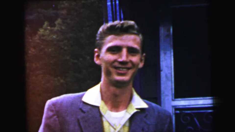 Young adult man in early-1960s narrow-lapel blazer and open-collar shirt smiling at camera, decorative iron gate and green shrub visible, circa 1964.