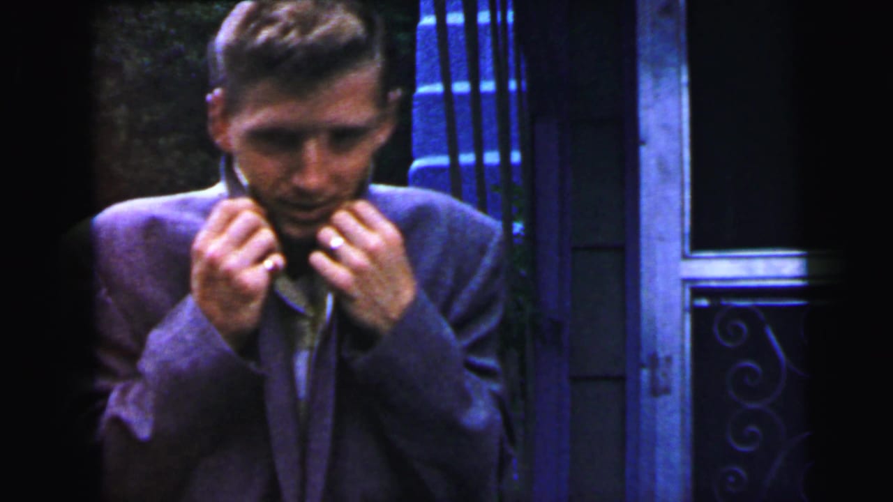 Same young man adjusting his collar with both hands raised, decorative ironwork and window visible at right, early-1960s suburban exterior, circa 1964.