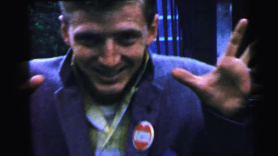 Close-up of young man grinning and raising fingers toward camera, a red circular lapel badge of unreadable text on his blazer, ornamental ironwork behind, circa 1964.