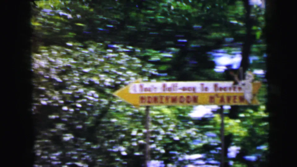 Yellow arrow-shaped roadside novelty sign with stacked text — possibly reading "Honeymoon Haven" — photographed on 8mm in woodland near Camden, New Jersey, 1964.