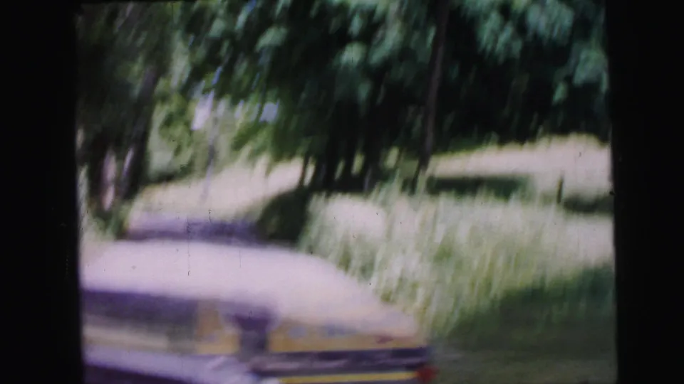8mm home-movie frame circa 1964 showing a wooded lane with a partially visible yellow sign at frame bottom, heavy motion blur from moving camera.