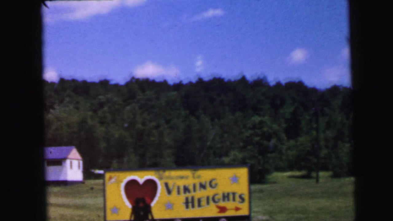 8mm home movie circa 1964: Viking Heights Resort entrance billboard with red heart graphic approached from a moving vehicle; open field and deciduous forest visible in background.