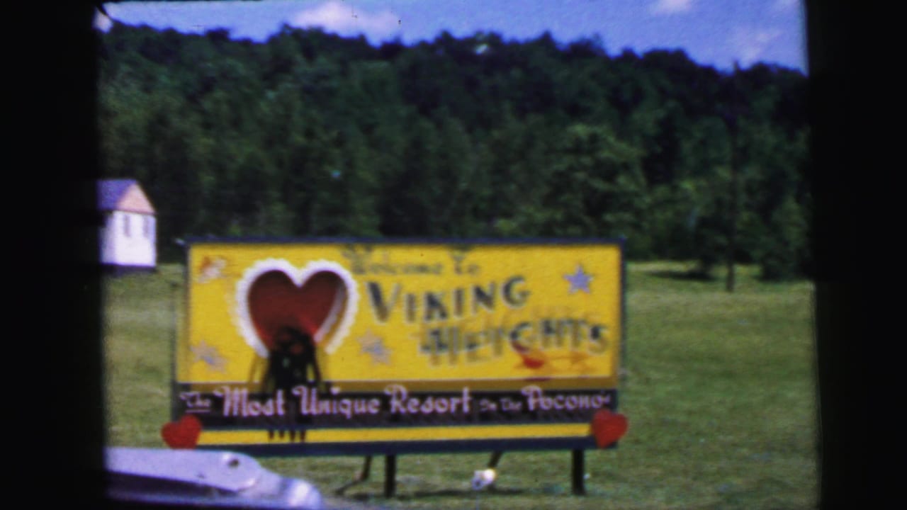 8mm footage circa 1964: Viking Heights Resort welcome billboard at closer range, full sub-banner 'The Most Unique Resort In the Poconos' legible; white outbuilding and forest in background.