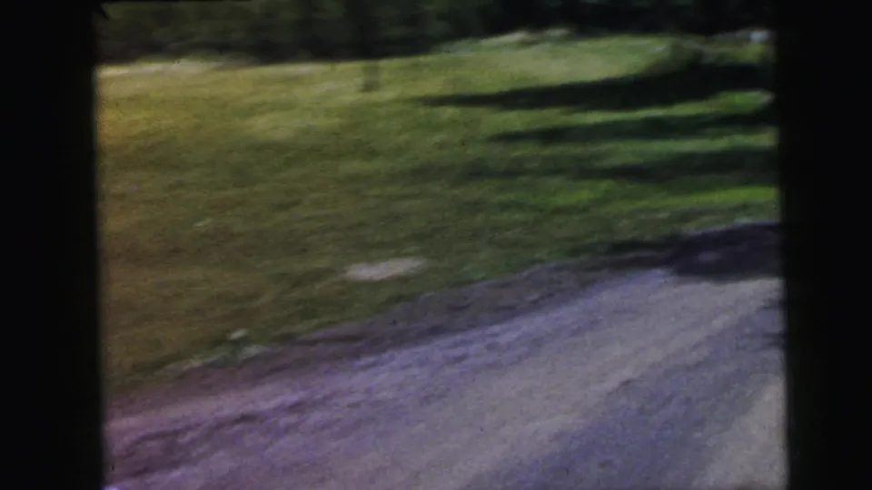 8mm home movie circa 1964: low passing-shot frame showing grass and road surface from inside a moving vehicle near the rural resort approach; minimal detail visible.