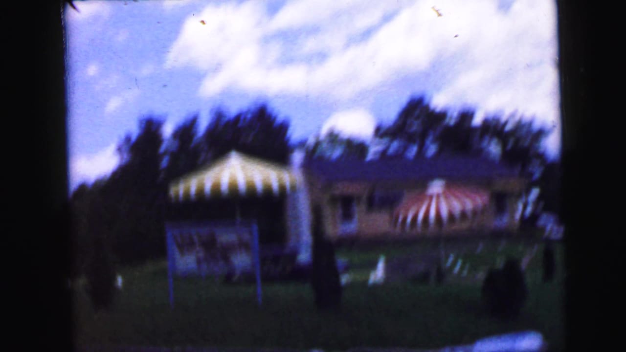 8mm home movie frame, circa 1964, showing a suburban backyard with two striped canopy structures and a low house, attributed to Camden, New Jersey.