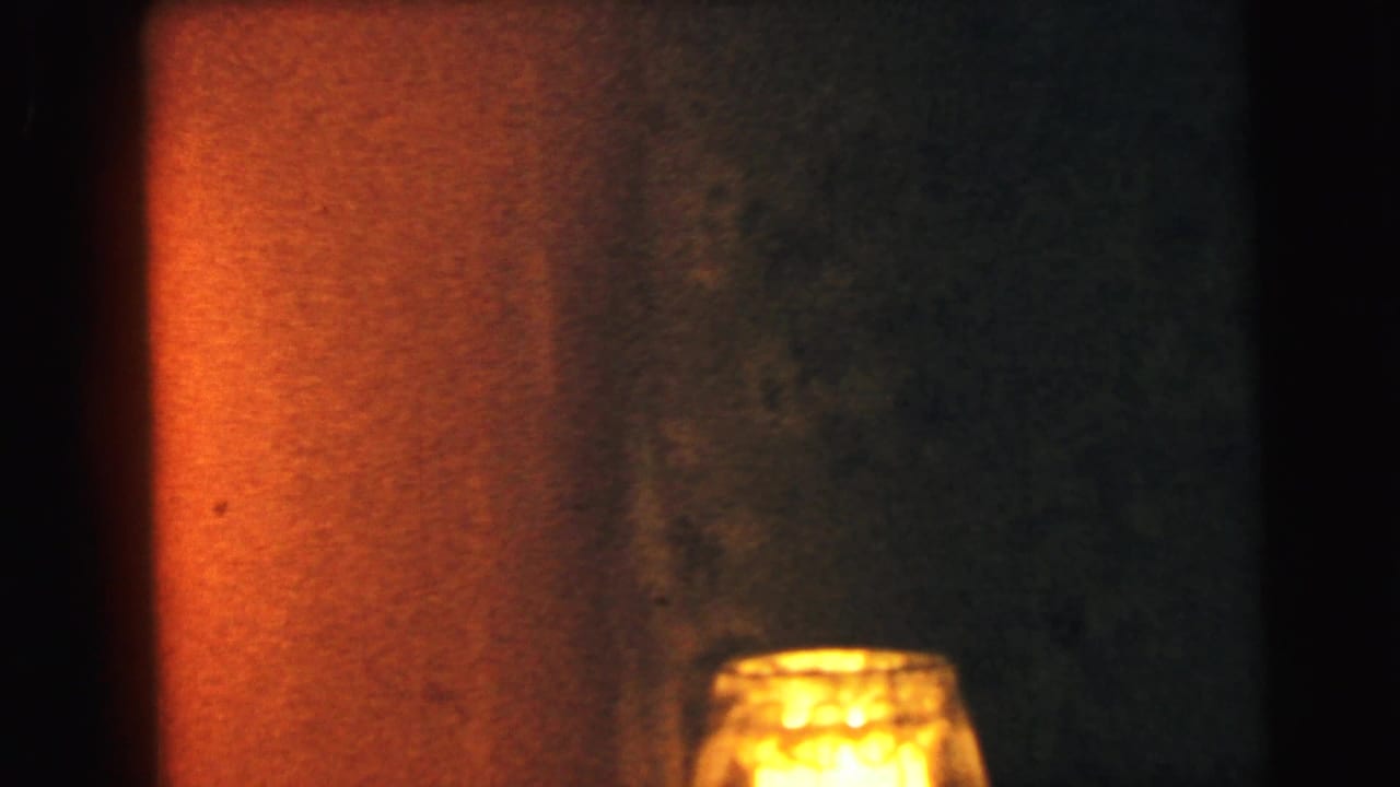 Close-up of a small illuminated glass lamp against an amber-lit wall, from an 8mm home movie in Camden, New Jersey, circa 1964.