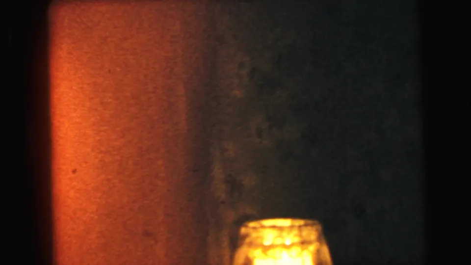 Close-up of a small illuminated glass lamp against an amber-lit wall, from an 8mm home movie in Camden, New Jersey, circa 1964.