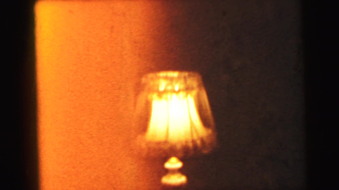 A 1960s-style table lamp with a pleated fabric shade on a pedestal base, illuminated against an orange-amber wall, Camden, New Jersey, 1964.