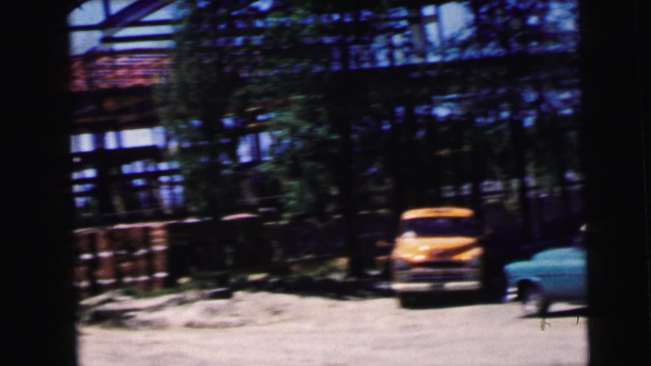 Yellow vintage vehicle and turquoise sedan parked on a dirt driveway beside an exposed structural frame, trees behind.