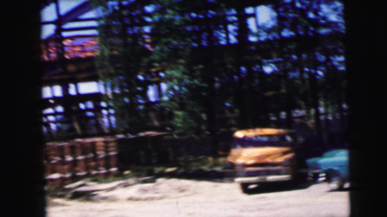 Yellow vintage vehicle centered on a dirt lot with a turquoise car to the right and exposed construction framing in the background.