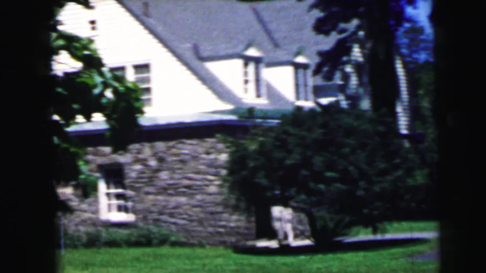 1964 8mm home movie frame: stone-and-siding residential house with gray roof, green lawn, and leafy shrubs, likely in New Jersey.