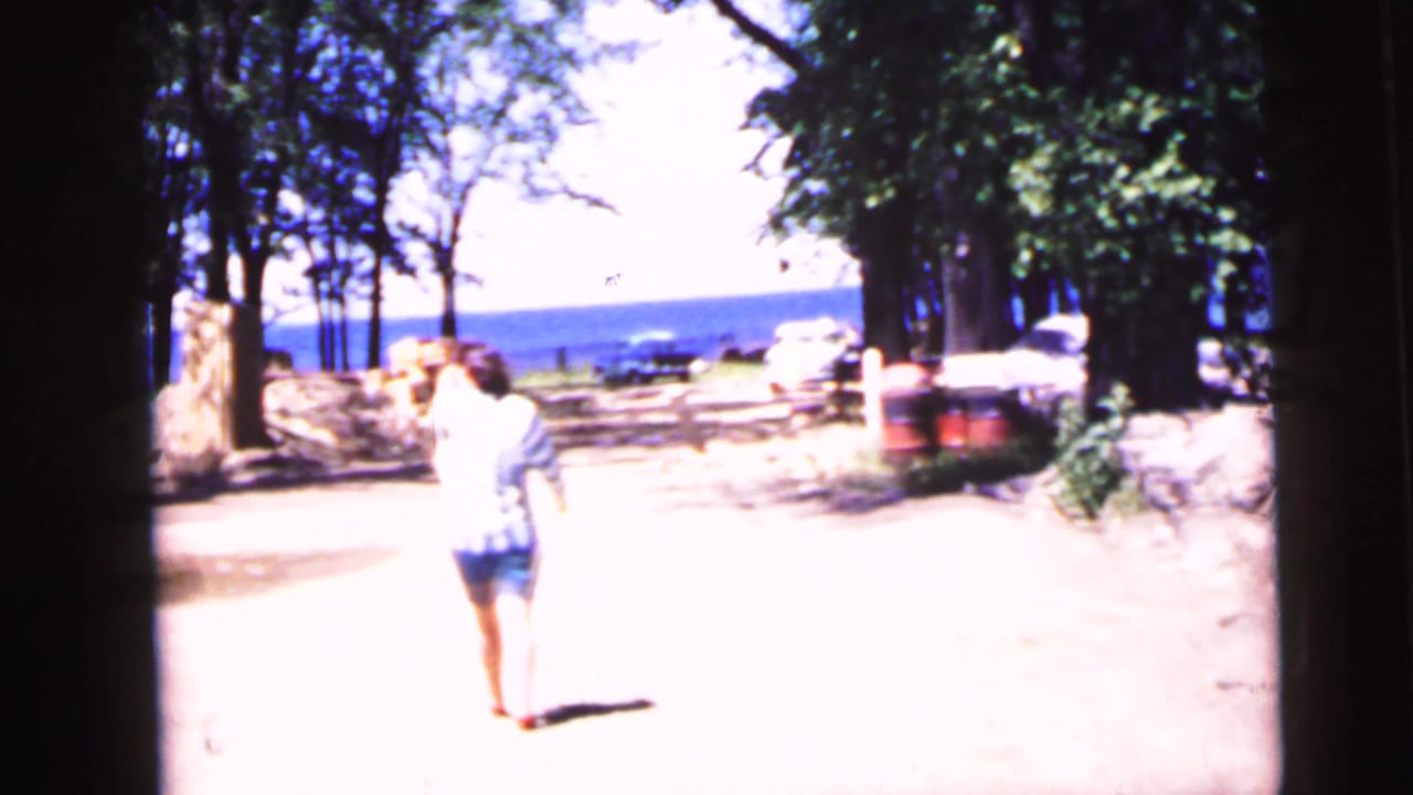 Woman in striped shirt and shorts walking on a sandy path near water, wooden fence and trees visible behind her.