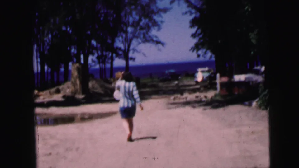 Woman in striped shirt and shorts walks away from camera along a sandy path toward a blue lake, flanked by trees and tree stumps; 1964 8mm home movie.