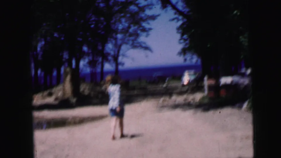 Woman in striped shirt and shorts continues along a sandy path toward a blue lake, trees framing the scene on both sides; 1964 8mm home movie.