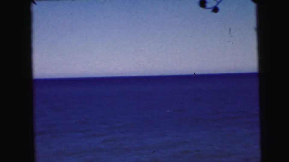8mm home movie frame, circa 1964, showing a panoramic view of blue-purple water under a pale sky with film dust artifacts and grain visible throughout.
