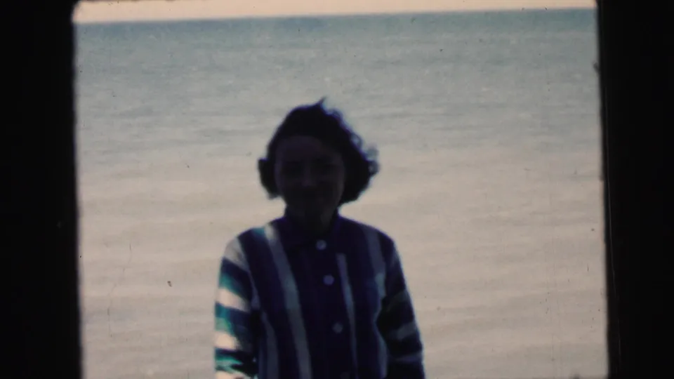 A woman in a 1960s-style striped cardigan poses in front of a large body of water, captured on 8mm film during what appears to be a family vacation circa 1964.