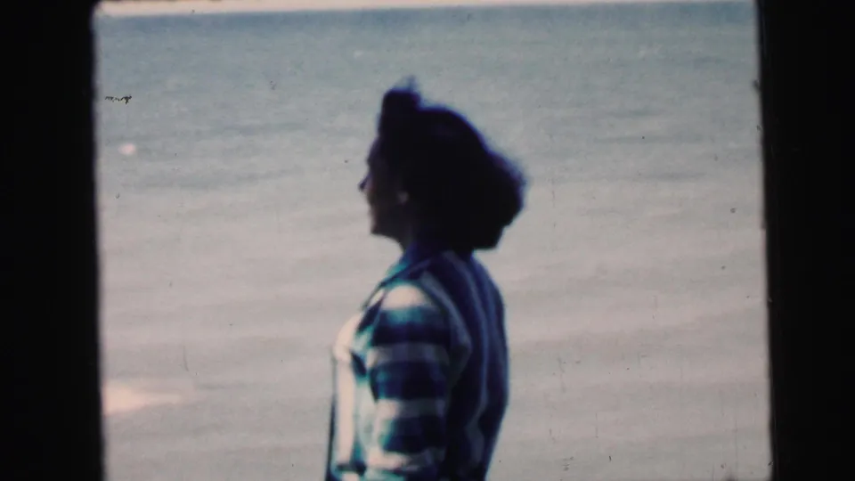 Woman in striped knit sweater in left profile, appearing to move along the shoreline with open water behind her, 8mm home-movie footage circa 1964.
