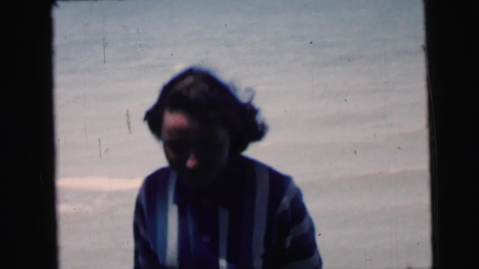 Woman in striped sweater glancing downward with hair slightly wind-blown and face partially visible, 8mm home-movie footage circa 1964.