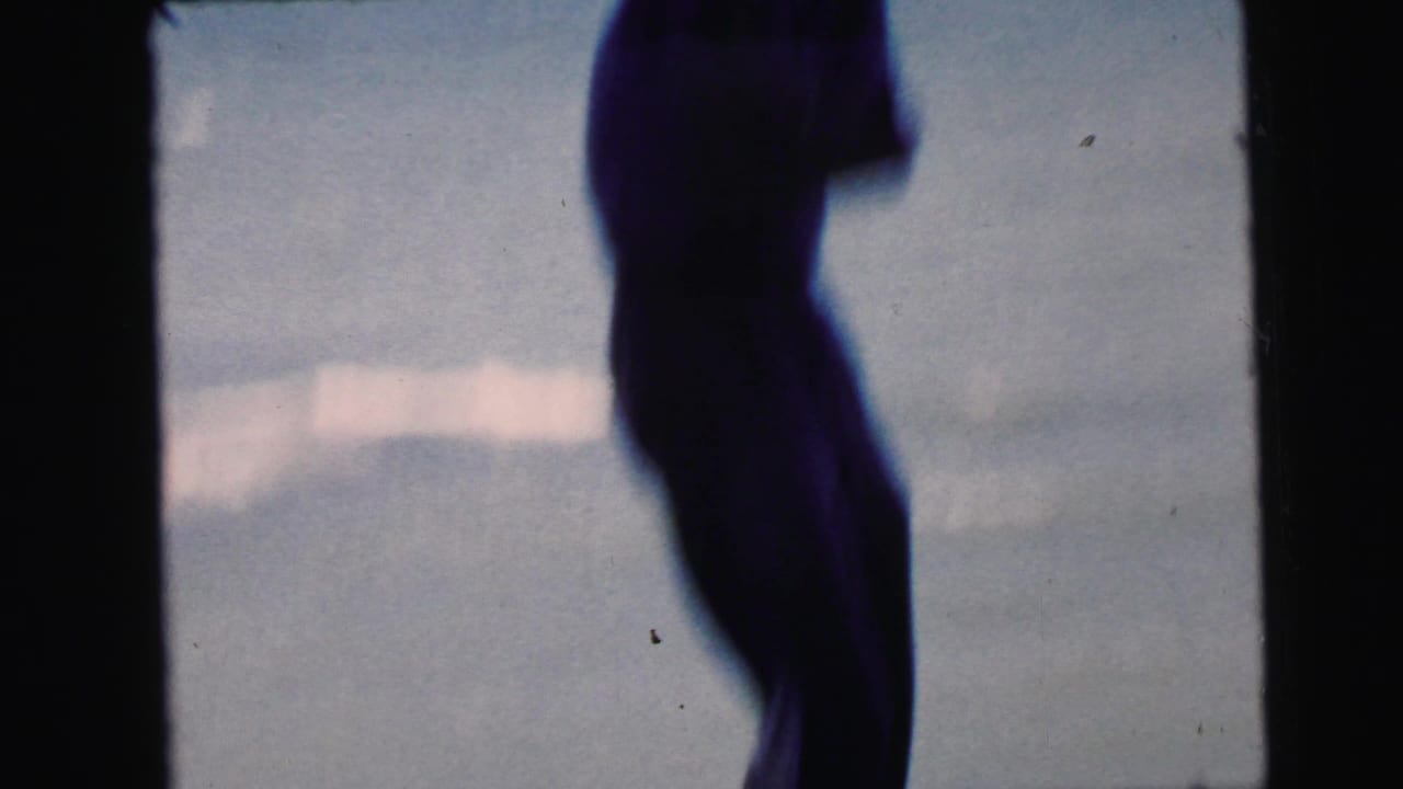 Near-total silhouette of a standing person against bright blue water and hazy sky, 8mm home movie footage, circa 1964, Camden New Jersey waterfront.