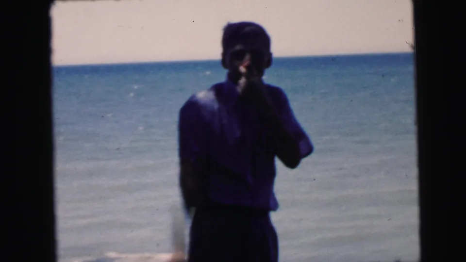 Adult figure in dark blue top at a waterfront with hand raised near face, 8mm home movie film, circa 1964, possibly Camden New Jersey.